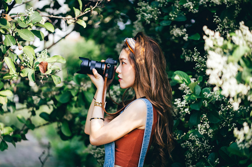 Side profile of beautiful Asian female university student photographing nature with camera in park on a sunny day
