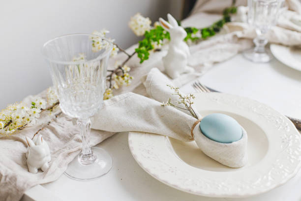 istock Stylish elegant Easter brunch table setting. Easter egg in bunny napkin on plate with cutlery, bunny, spring flowers on rustic white table. Modern Easter table arrangement 1371226129