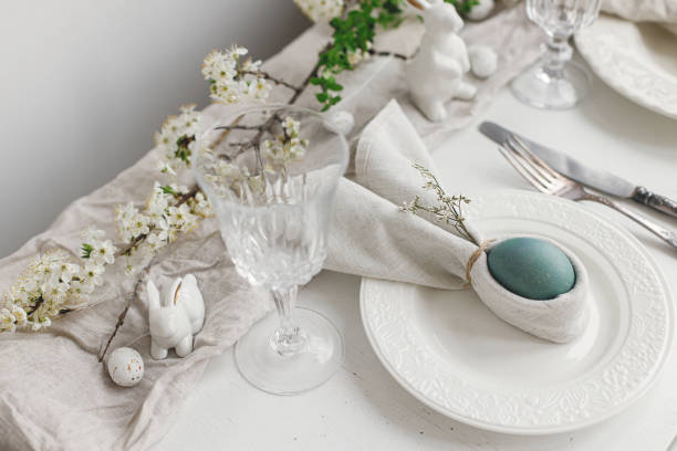 istock Stylish elegant Easter brunch table setting. Easter egg in bunny napkin on plate with cutlery, bunny, spring flowers on rustic white table. Modern Easter table arrangement 1371226732