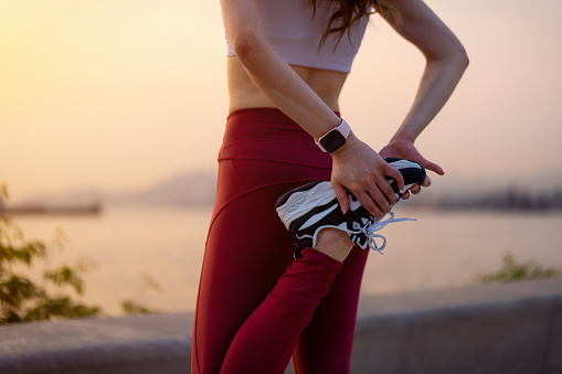 Cropped shot of young Asian sports woman in sportswear, stretching legs outdoors, standing by the promenade at sunset. Getting ready to exercise. Girl power. Health and fitness concept