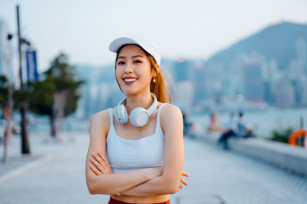 カメラを見て、腕を組んだ自信に満ちた若いアジアのスポーツウーマンの肖像画。彼女は市内の遊歩道のそばに屋外に立っているヘッドフォン付きのキャップを着用しています。若者の文化� - self improvement audio ストックフォトと画像