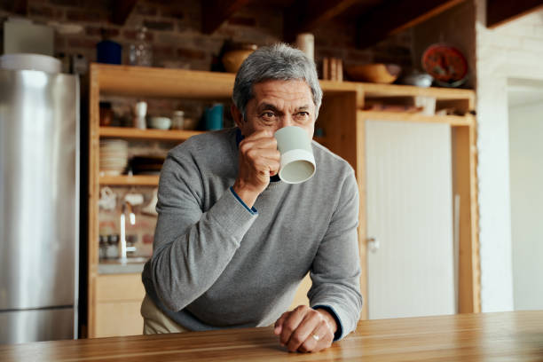 portrait of retired biracial elderly male leaning on modern kitchen counter, sipping on his morning coffee - senior adult human face male action imagens e fotografias de stock