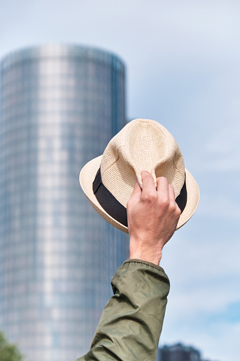 Man's hat in hand against the backdrop of a skyscraper office building and blue sky. Welcome and greeting hand gesture, goodbye office and freedom concept.