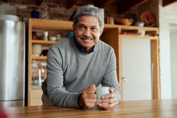 retrato de idoso biracial aposentado homem inclinado em balcão de cozinha sorrindo para a câmera - senior adult human face male action - fotografias e filmes do acervo