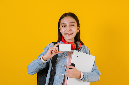 Young latin teen child girl student holding a blank empty card isolated on yellow background in Latin America