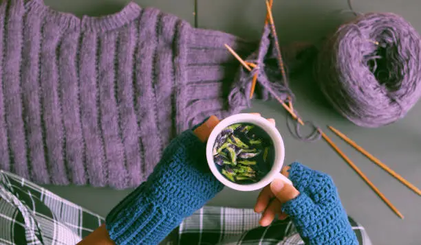 Top view woman hand wear blue mitten crochet, hold cup of tea while knit violet sweater for gift on dark tone