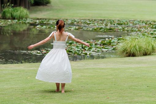 Portrait of young Asian woman dancing on grass field with barefoot in forest park, beautiful Chinese girl in white dress enjoy her carefree time in sunny summer day, full length shot rear view.