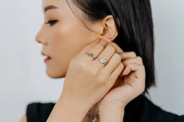 An Asian woman portrait is wearing diamond earring, looking at side down with white background. Portrait of beautiful Asian woman dressed in a posh jewelry set of diamond ring, bracelet and earrings