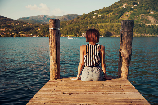 Redhead young woman back portrait sitting on wooden dock in front of Lake Orta, Italy