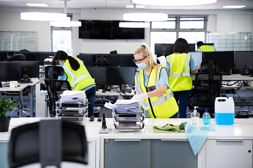 Caucasian woman and colleagues wearing hi vis vests, gloves, safety glasses and face masks sanitizing an office using disinfectant. Hygiene in workplace during Coronavirus Covid 19 pandemic.