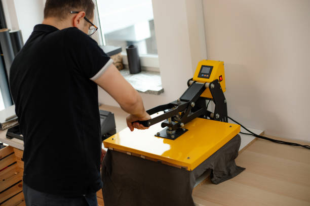 Man worker glasses back using heat thermal textile application transfer machine. Printing application art design T-shirt Man worker in glasses back using heat thermal textile application transfer machine. Printing application art design black T-shirt. Printing house office offset equipment. Manufacturing business label t shirt transfer stock pictures, royalty-free photos & images