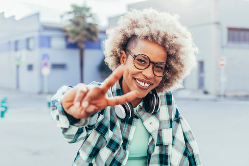 Young latin american afro millennial woman with curly hair listening to music, using white headphones smiling with happy face, looking at camera doing victory sign outdoors.