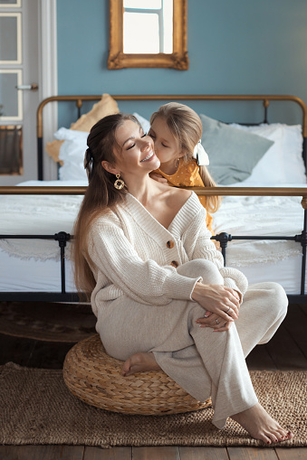 The concept of a happy relationship between mother and daughter. Tender baby girl kissing her beautiful mom, sitting on floor at domestic living room with modern retro interior. Happy mothers day.