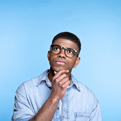 Portrait of pensive african young man wearing denim shirt and eyeglasses, looking up with hand on chin. Studio portrait on blue background.