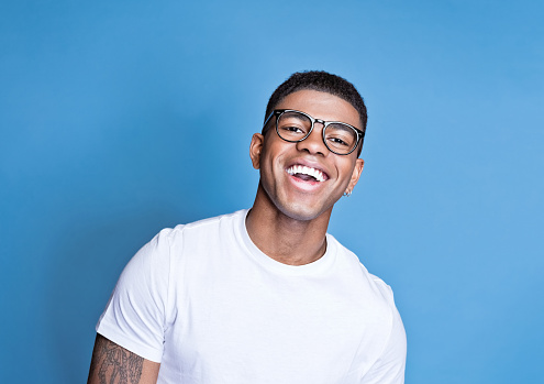 Happy african young man wearing white t-shirt and eyeglasses, laughing at camera. Studio portrait on blue background.