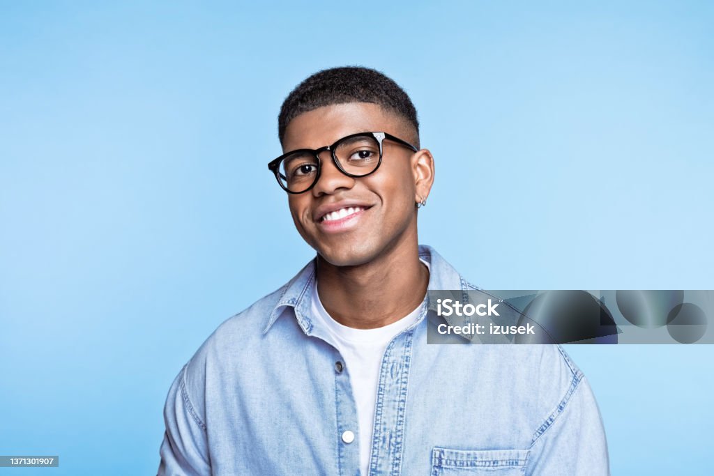 Friendly young man wearing denim shirt Confident african young man wearing denim shirt and eyeglasses, smiling at camera. Studio portrait on blue background. Men Stock Photo