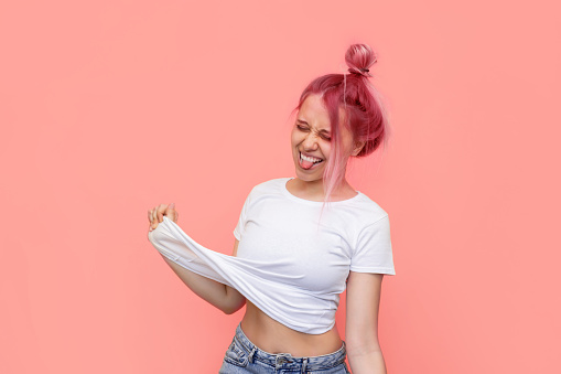 A young caucasian ridiculous woman with pink hair shows tongue grimacing and pulling up her white t-shirt on a light pink color background. Funny informal girl fooling around