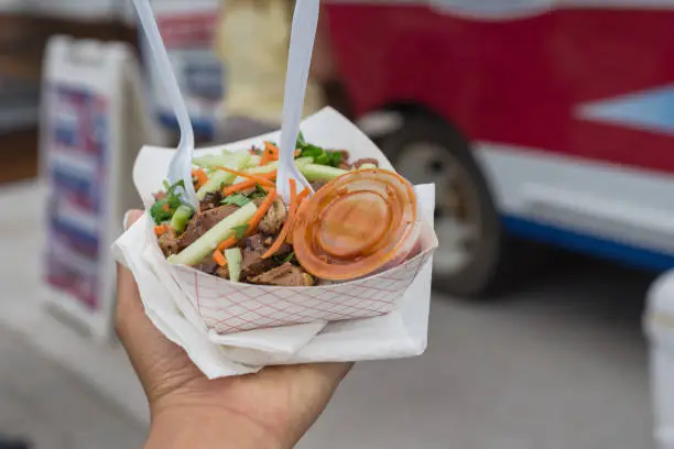 Take away paper food tray of chicken teriyaki with chili sauce in deli containers, napkins, fork and spoon with blurry food truck background. Togo grilled meat with allumette cut cucumber and carrot slice