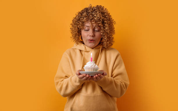 アフロカール髪型を持つ魅力的な若い30年代の女性の肖像画は、黄色の壁の背景の上に孤立したろうそくで誕生日ケーキを保持して喜んだ表情をしています - cake birthday candle blowing ストックフォトと画像