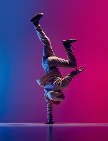 Studio shot of young flexible sportive man dancing breakdance in white outfit on gradient pink blue background. Concept of action, art, beauty, sport, youth. Dancer shows breakdance figures