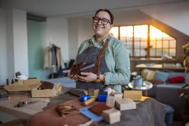 Portrait of mid adult woman in her leather crafting workshop, she's holding handmade wallets, looking at camera and smiling