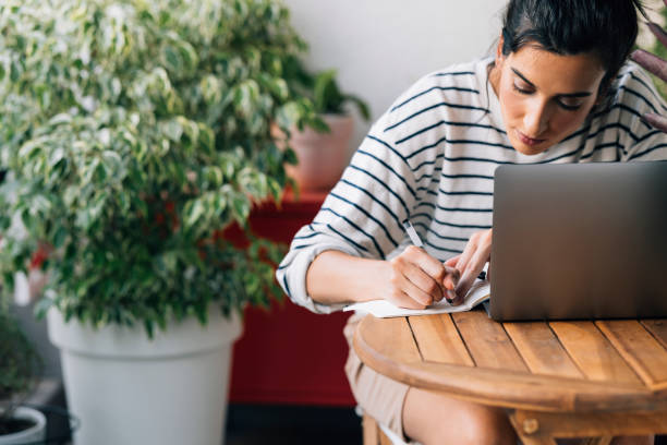istock Beautiful Woman Sitting Surrounded by Potted Plants, Writing Down Notes and Using Her Laptop 1371322979