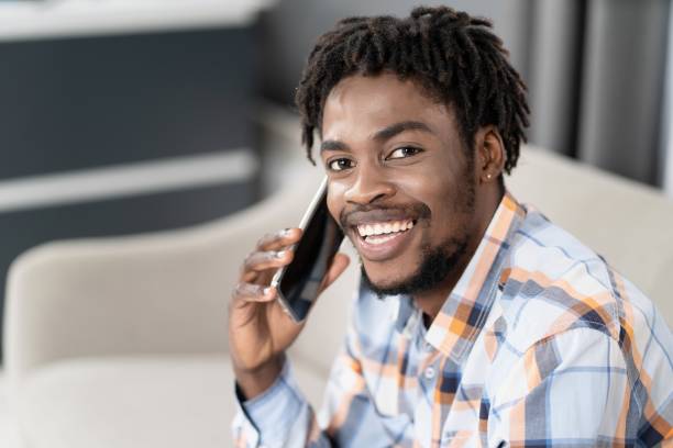 istock Young African American guy with phone in hand looking at camera while having conversation. Portrait of happy man having present conversation sitting on the sofa. Social media concept 1371328718