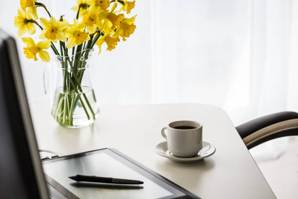 Selective focus shot of a digital tablet with a digital pen on it, a cup of coffee and a bouquet of yellow daffodils in a glass vase on a office desk.