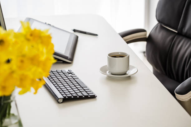 Shot of a office desk with a cup op coffee, digital tables, desktop computer and a computer keyboard with some yellow daffodils blurred in the foreground.