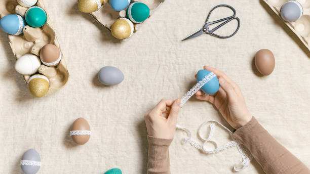 istock Decoration Easter eggs, female hands decorating colorful chicken egg with white tape lace, traditional celebration food and holiday symbol.  Home preparing for Happy Easter. Top view 1371341556