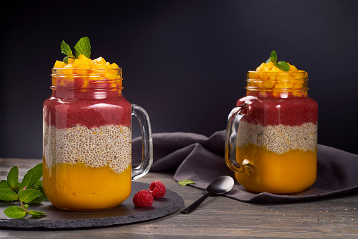 Two jars of layered chia pudding with mango and raspberries on dark wooden background