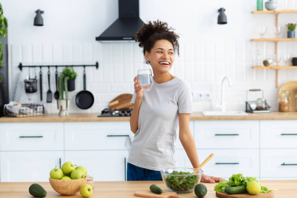 istock Healthy diet eating. African American young female preparing salad for dinner or breakfast in home kitchen. Beautiful woman cooking healthy food with glass of clean water, looking away, smiling friendly 1371347607