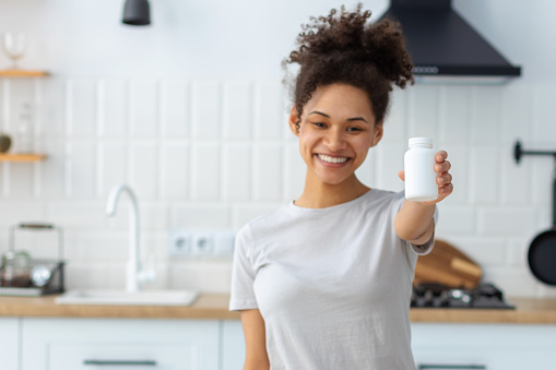 Happy young African American woman holding bottle of dietary supplements or vitamins in her hands. Close up. Healthy lifestyle concept