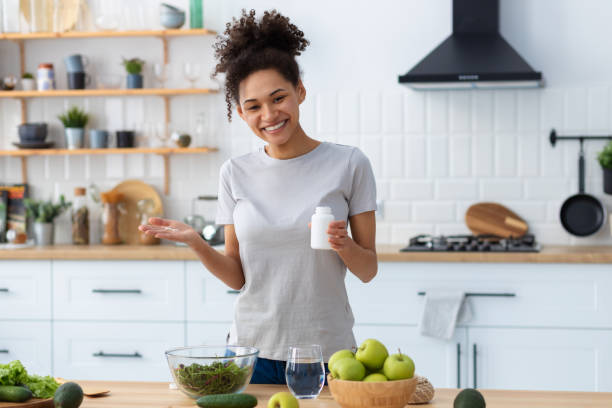 femme afro-américaine heureuse dans la cuisine de la maison tenant une bouteille de suppléments nutritionnels, regardant la caméra et souriant un mode de vie amical et sain - vitamin pill nutritional supplement capsule antioxidant photos et images de collection