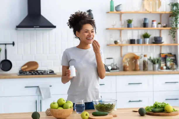 Photo of Happy african american woman standing at the cuisine table in the home kitchen drinking dietary supplements, looking away and smiling friendly, healthy lifestyle concept