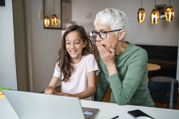 senior woman and a cute girl at home, looking at the laptop - wireless technology cheerful granddaughter grandmother imagens e fotografias de stock
