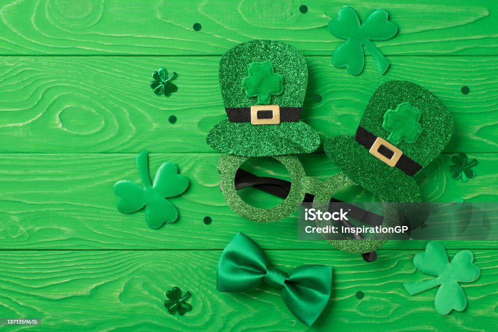Top view photo of st patrick's day decorations shamrocks trefoil shaped confetti funny outfit hat shaped party glasses and green bow-tie on isolated textured green wooden desk background St. Patrick's Day Stock Photo