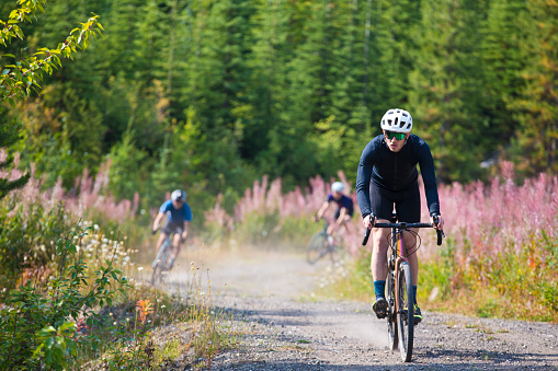 A man goes for a ride on his gravel bicycle in the Rocky Mountains of Canada. He is followed by two other riders in the background. Gravel bikes are similar to cyclo-cross bicycles with sturdy wheels and tires for riding on rough terrain. His bike has a large frame bag to carry extra gear.