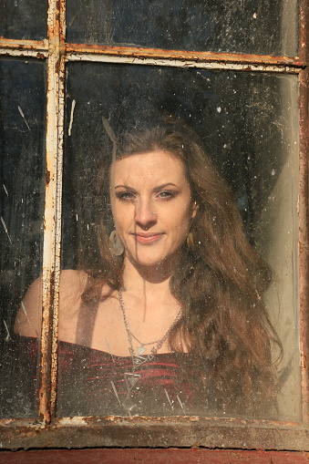 A woman looking through a dirty window. She is wearing a red off shoulder velvet top and  a necklace with earrings.