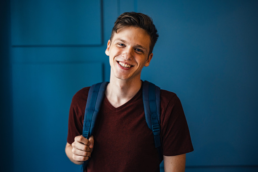 Portrait of a young male standing, smiling and wearing a backpack. There is a blue background behind him.