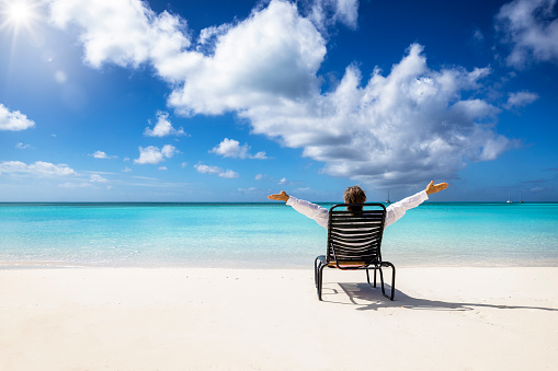 A happy man relaxes in a chair on a tropical paradise beach in the Caribbean with turquoise sea and blue sky during his vacation time
