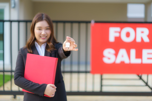 Asian real estate agent or Real Estate Agent woman smiling and holding red file with showing house key in front of house.