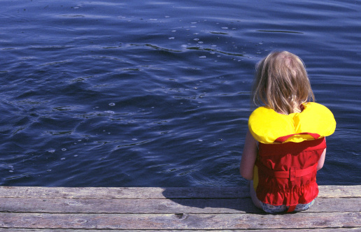 A young, blonde, girl in a bright red and yellow life jacket sits on a wooden dock and creates ripples and bubbles by dipping her toes in the cool, blue water of a mountain lake.