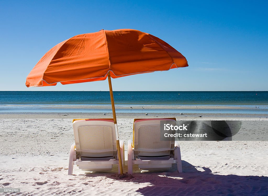 A setting on the beach of beach chairs and an umbrella  A bright orange beach umbrella and yellow cushioned beach chairs await sunbathers along the beach at Fort Myers Beach, Florida. Beach Umbrella Stock Photo