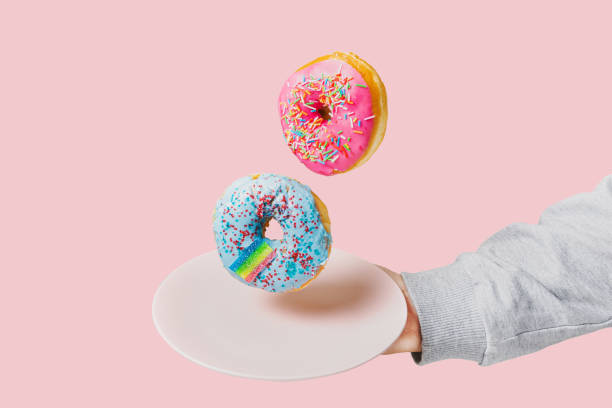 istock Two multi-colored donuts hover over a plate held by a hand on a pink background. Unhealthy food concept 1371428183
