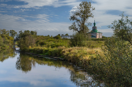 Summer landscape in the city limits of Solikamsk (Russia). The raised bank of the river, overgrown with grass, trees with kutarniks, which are reflected in the water. In the distance the old church