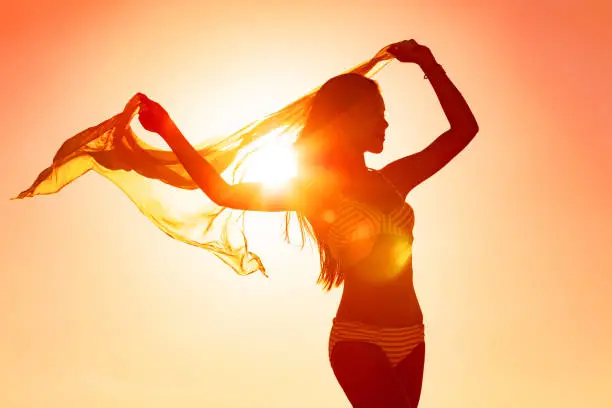 Photo of Wellness woman body silhouette curves dancing carefree with femininity with scarf flowing in the wind at sunset glow flare for life happiness. Sun vacation travel beach