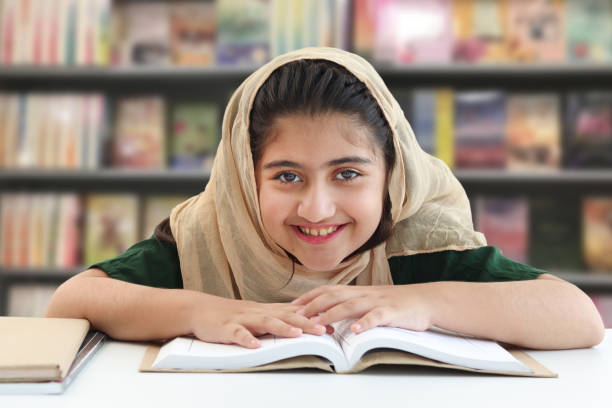 adorabile ragazza musulmana pakistana sorridente con bellissimi occhi che indossano l'hijab, studiano e fanno i compiti sul tavolo, bambino studente felice che legge il libro sullo sfondo sfocato della libreria in biblioteca. - islam child indian culture ethnic foto e immagini stock