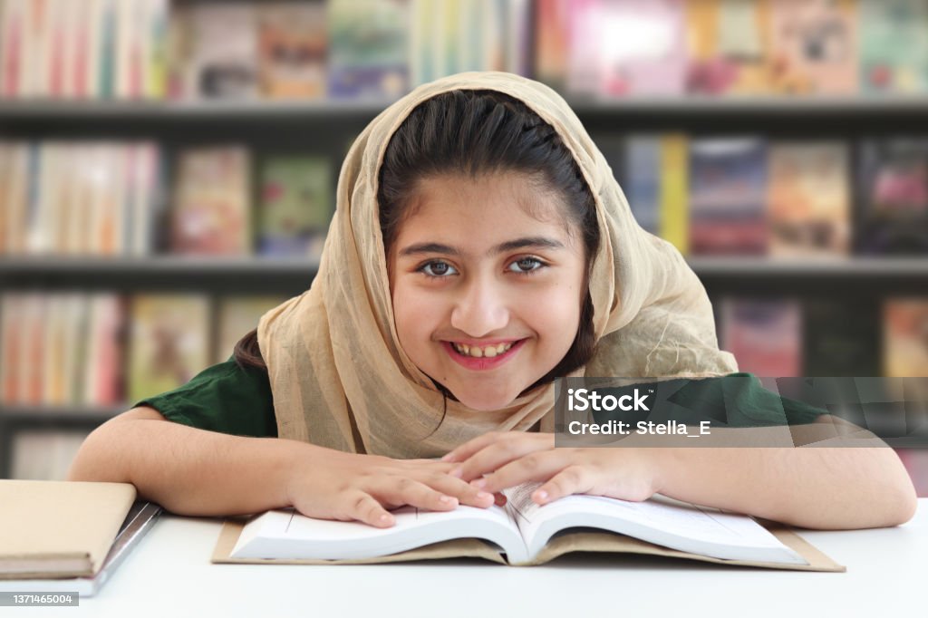 Adorable smiling Pakistani Muslim girl with beautiful eyes wearing hijab, studying and doing homework on table, happy student kid reading book on blurred background of bookcase in library. Child Stock Photo