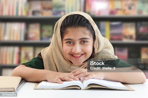 Adorable Smiling Pakistani Muslim Girl With Beautiful Eyes Wearing Hijab Studying And Doing Homework On Table Happy Student Kid Reading Book On Blurred Background Of Bookcase In Library Stock Photo - Download Image Now